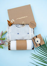 Load image into Gallery viewer, An image of a Baby girl gift hamper with Indigo cotton romper and swaddle, alomg with a hair accessory and a toy, on a light bluish background.