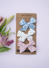Load image into Gallery viewer, A zoomed out picture of three elegant knot combo hairclips for girls, one in white colour with small black dots, the other in a pinkish flowery print, and a third with light blue floral print, on a light blue background.