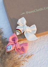Load image into Gallery viewer, Two Hair Clips with glitters are sitting on a white surface next to flowers and another has paste colored bows with a rainbow