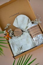 Load image into Gallery viewer, A beautiful gift box filled with unisex newborn gift hamper with swaddle made of wild silver oak with a toy and some flowers inside the box which is kept upon a pink background with some leaves aside.