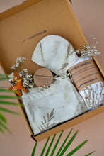 Load image into Gallery viewer, A beautiful gift box filled with unisex newborn gift hamper with swaddle made of wild silver oak with a toy and some flowers inside the box which is kept upon a pink background with some leaves aside.