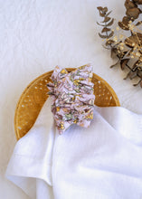 Load image into Gallery viewer, A hair accessory of Lavender Floral Scrunchies placed in a wooden basket with a white cloth beneath, placed on a white background with some leaves and decoratives