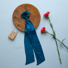 Load image into Gallery viewer, A beautiful blue silk hair scrunchie kept upon a round wooden log with some flowers and brown card aside.