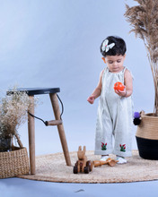 Load image into Gallery viewer, A baby girl wearing cute jumpsuit with matching hair accessories with some artificial plants and wooden pots and with some rabbit toys and a flower basket aside.