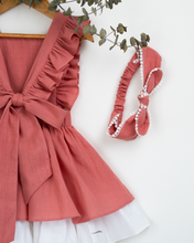 Load image into Gallery viewer, A back view of a beautiful dress with a criss-cross back hung on a hanger with a matching hair accessory beside it with a stem of leaves upon it.