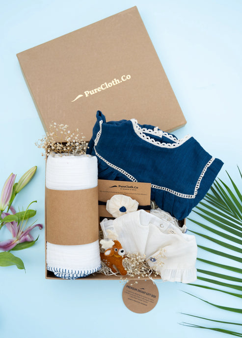 An image of a Baby girl gift hamper with Indigo cotton romper and swaddle, alomg with a hair accessory and a toy, on a light bluish background.