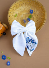 Load image into Gallery viewer, A hair accessory of White Hair Bows - Blue Floral placed in a wooden basket, placed on a light brown background with some tiny blue flowers, leaves and decoratives
