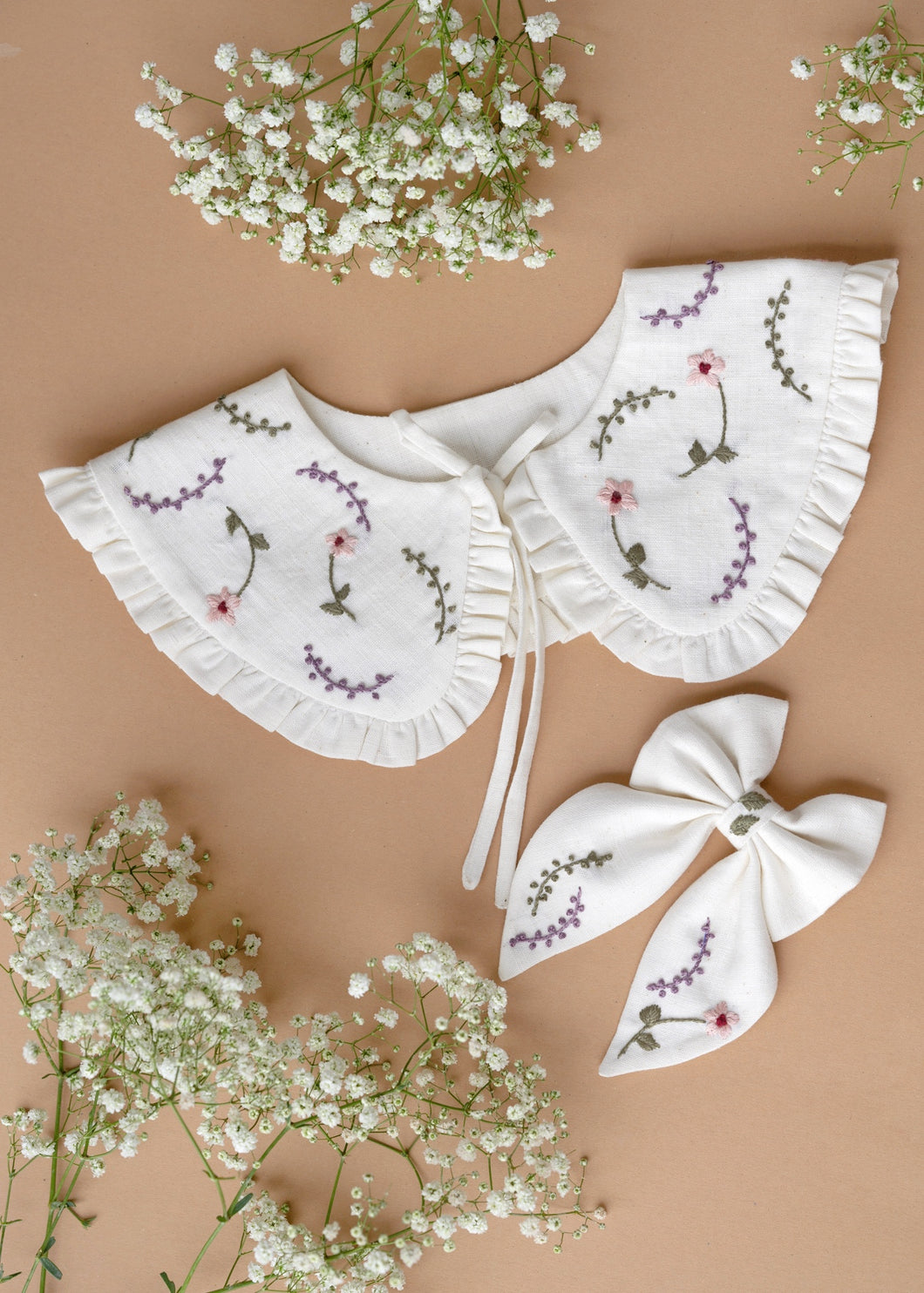 A beautiful combo of detachable victorian style collar and matching bow with subtle hand embroidery for kids kept on light peach background with some flowers aside.