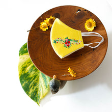 Load image into Gallery viewer, A beautiful organic embroidery face mask kept on a wooden plate with some flowers and leaf aside.
